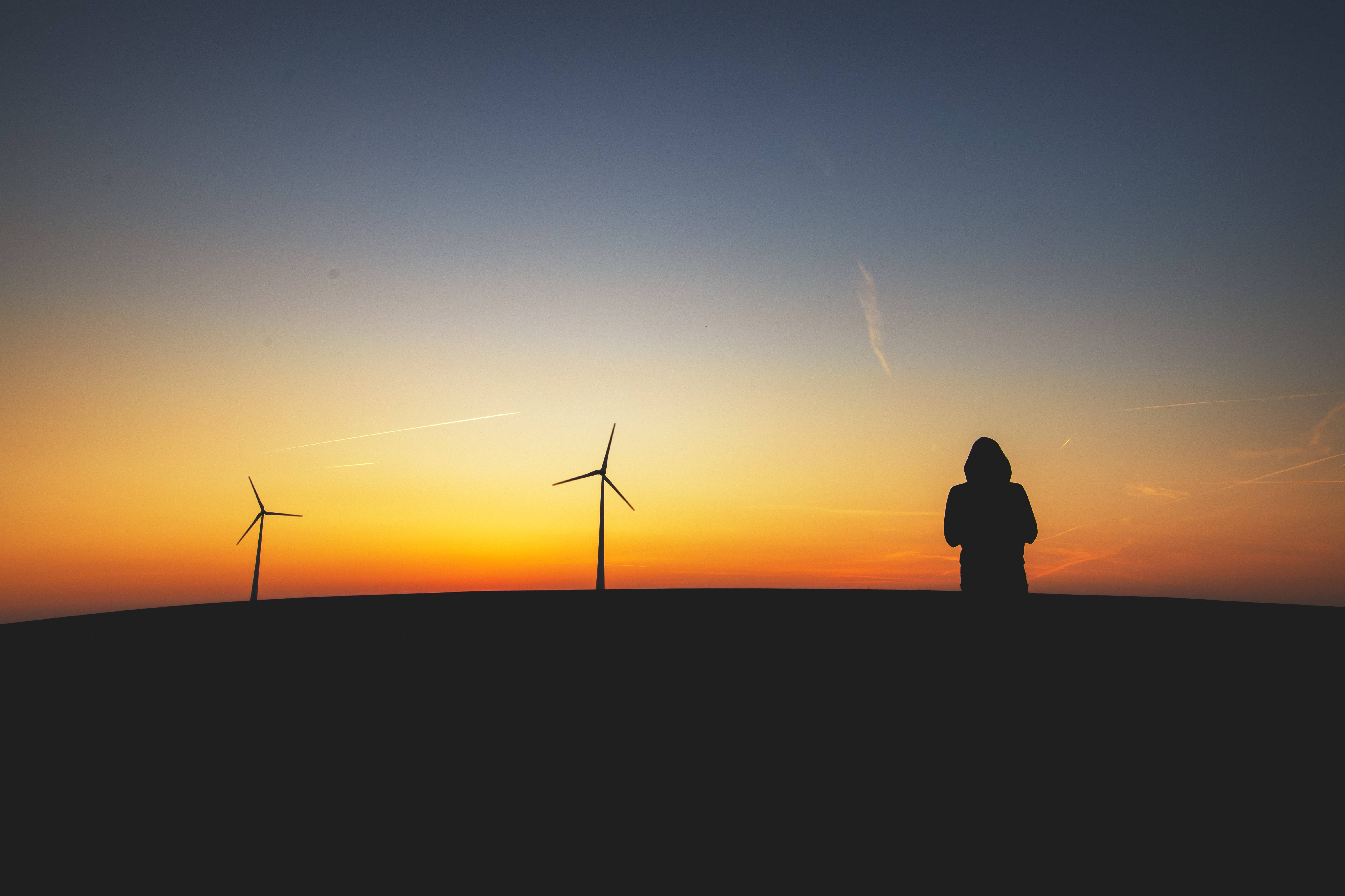 A person stands with windmills and the sunset in the distance.